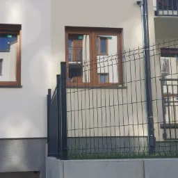 Kamera monitoringu zamontowana na elewacji budynku obok rury spustowej, widoczna przez metalowe ogrodzenie balkonu.