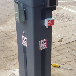 P.H.U ANDRZEJ GRZESIAK - Szara kolumna z kamerą, przyciskiem SOS i naklejkami ostrzegającymi o całodobowym monitoringu, na kostce brukowej, obok leży śrubokręt.