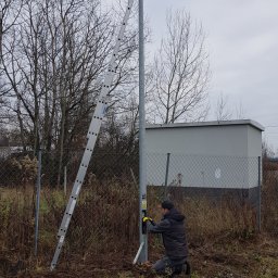 P.H.U ANDRZEJ GRZESIAK - Instalator w żółtych rękawicach klęczy przy słupie z kamerą przemysłową, podłączając przewody; drabina oparta o słup, w tle szara budka i gołe drzewa.