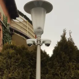 Lampa uliczna z dwoma kamerami kopułkowymi Dahua, otoczona tujami, na tle budynku z balkonem.