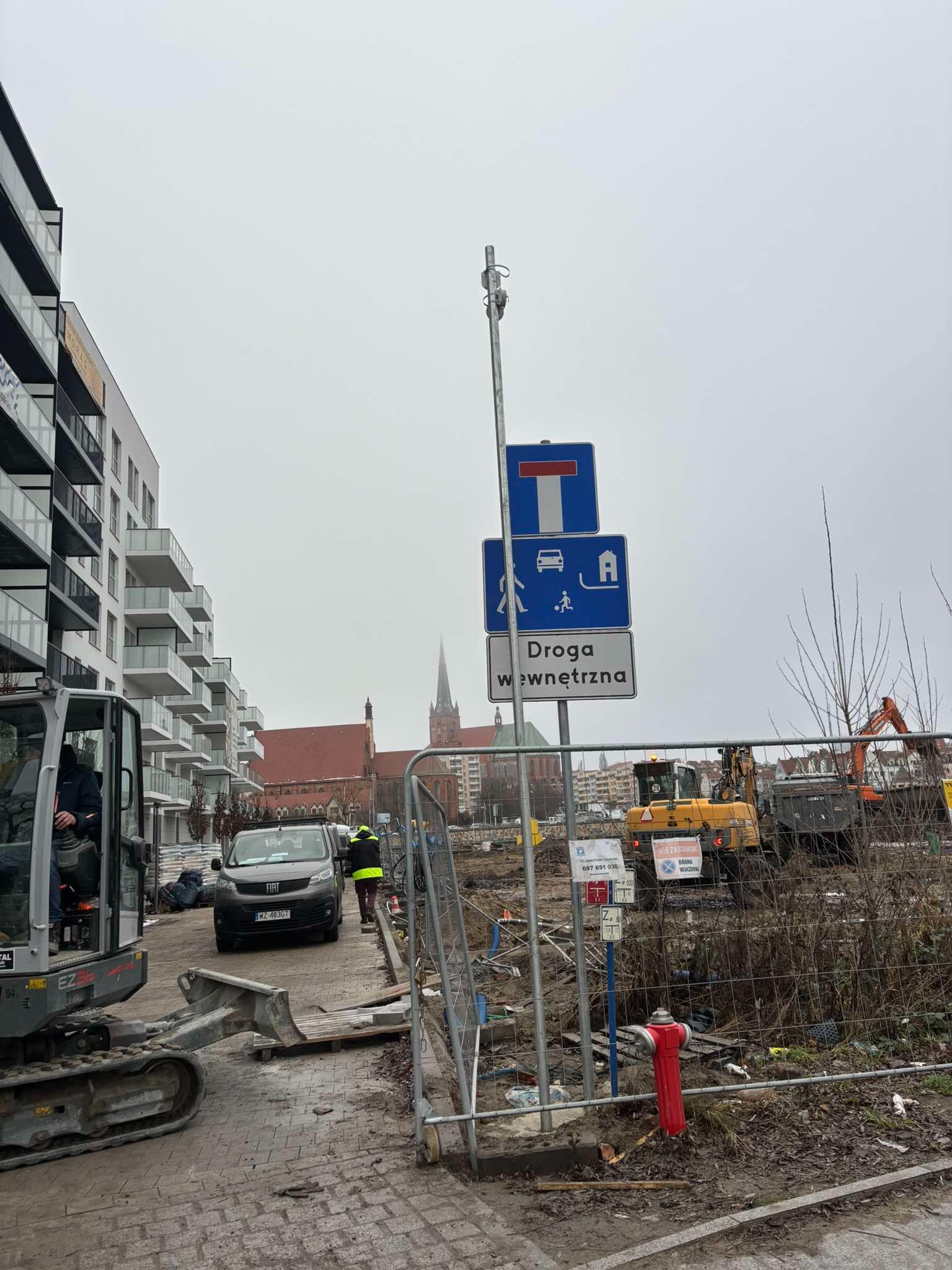 Teren budowy z widocznym sprzętem ciężkim, znakiem drogowym 'Droga wewnętrzna' oraz nowym budynkiem mieszkalnym w tle, uchwycone w pochmurny dzień.