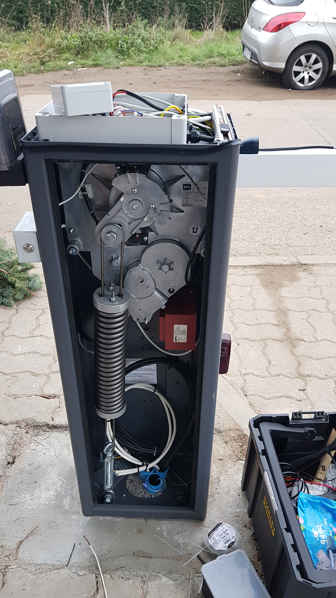 Otwarta obudowa automatycznej bariery parkingowej z widocznym mechanizmem sprężynowym, przekładniami i okablowaniem; skrzynka narzędziowa Stanley obok.