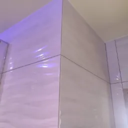 Ściana narożna wyłożona jasnymi, marmurkowymi płytkami ceramicznymi, z widocznymi fugami w kolorze szarym, oświetlona fioletowym światłem.