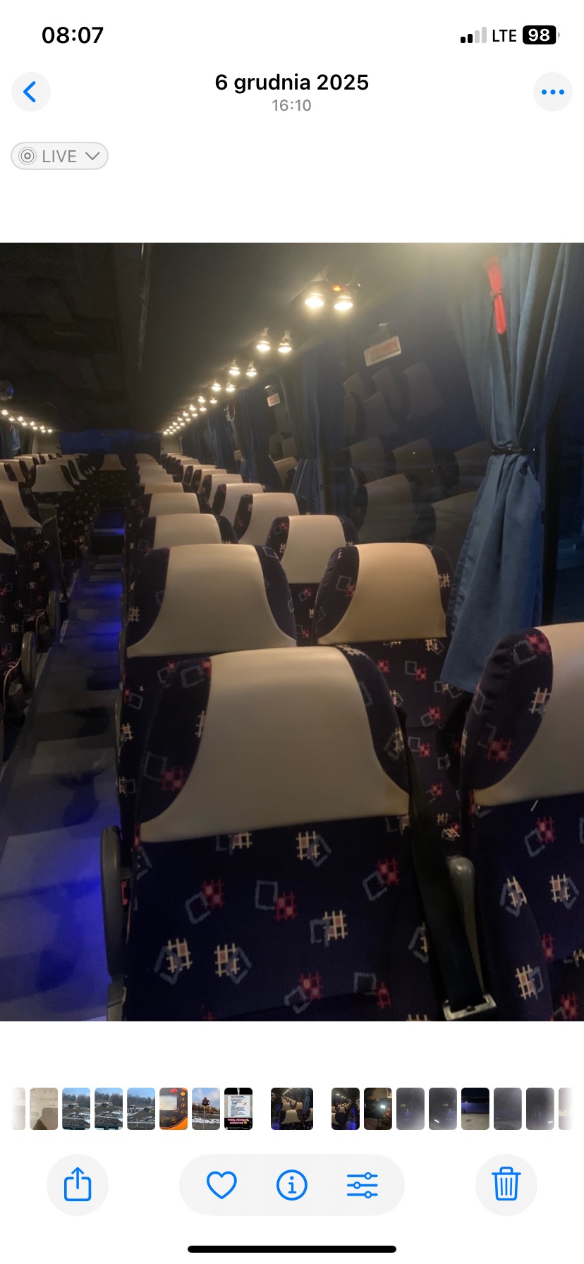 Wnętrze autobusu z rzędami foteli z białymi zagłówkami i wzorzystą tapicerką. Widoczne oświetlenie sufitowe i zasłony przy oknach. Perspektywa od przodu pojazdu.