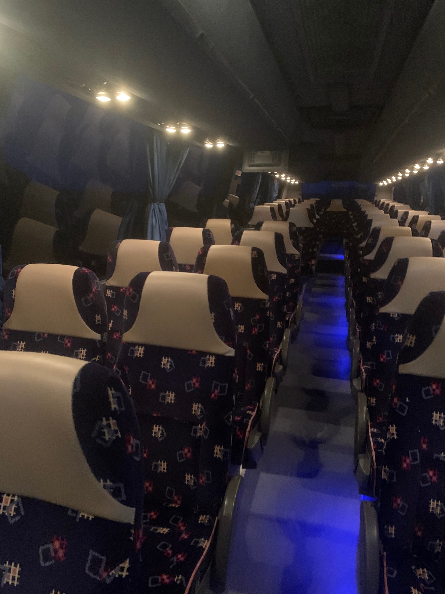 Wnętrze autobusu z rzędami foteli obitych wzorzystą, granatową tkaniną i beżowymi zagłówkami, oświetlone niebieskim światłem podłogowym i lampkami sufitowymi.