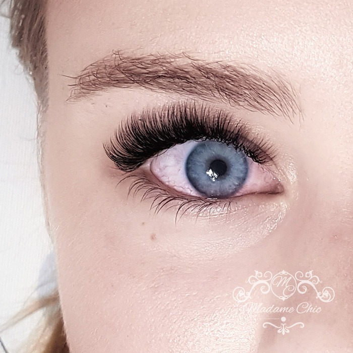 Zbliżenie na oko z niebieską tęczówką i wyraźnie przedłużonymi, gęstymi rzęsami; widoczny pieprzyk na policzku i logo w prawym dolnym rogu.