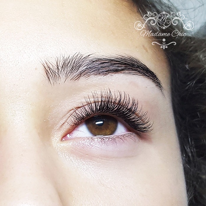Zbliżenie na oko z brązową tęczówką i gęstymi, czarnymi, przedłużonymi rzęsami. Widoczny delikatny makijaż oka i logo 'Madame Chic' w prawym górnym rogu.