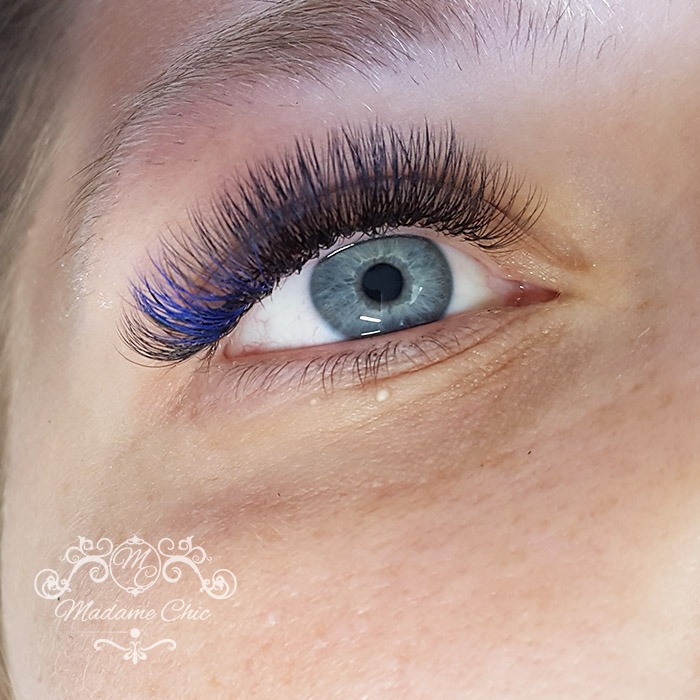 Zbliżenie na oko z niebieskimi akcentami w przedłużonych rzęsach, widoczne logo Madame Chic w lewym dolnym rogu.