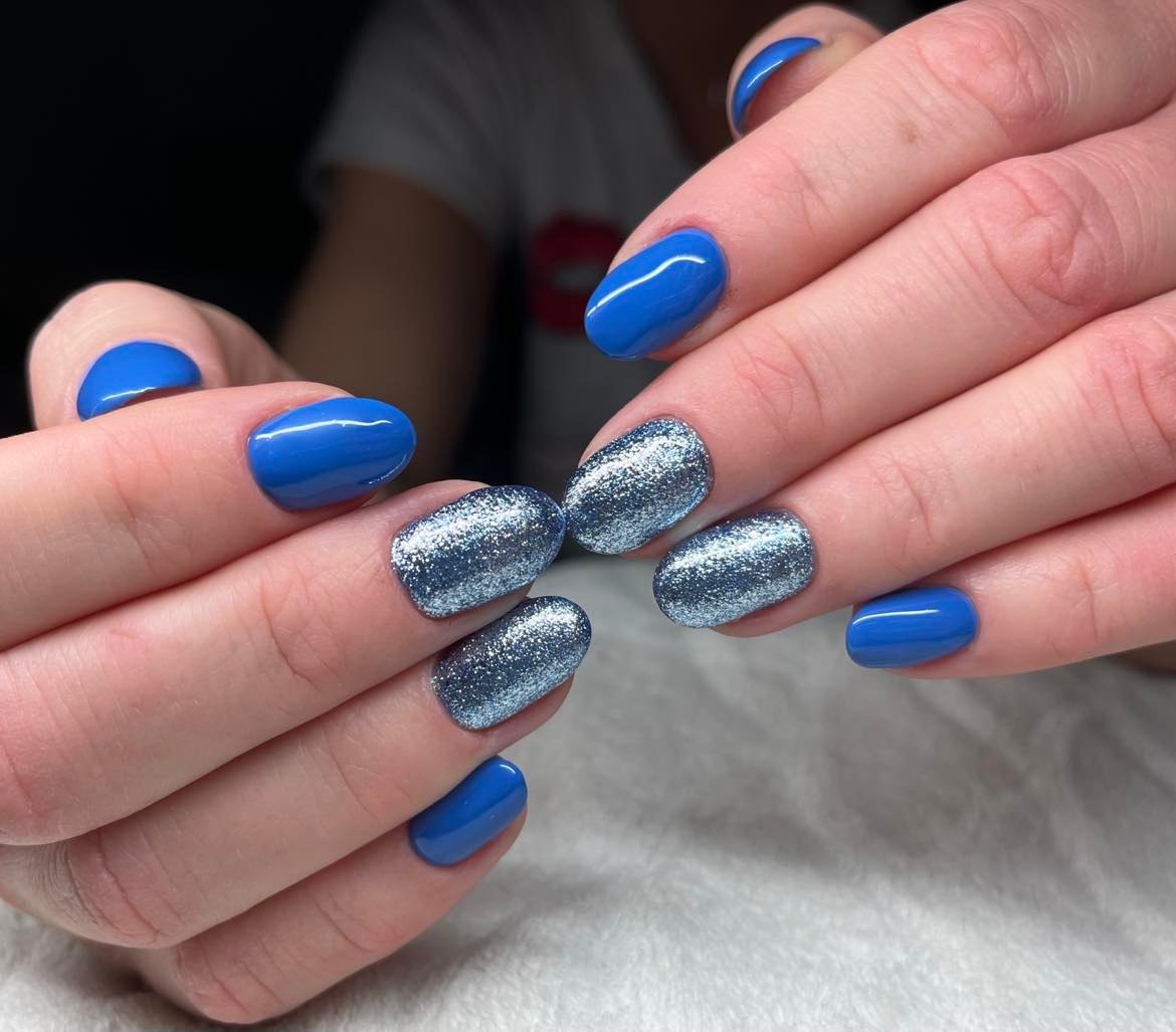 Manicure hybrydowy: kobaltowe paznokcie z brokatowym akcentem na serdecznym i środkowym palcu, owalny kształt, widoczne delikatne linie papilarne.