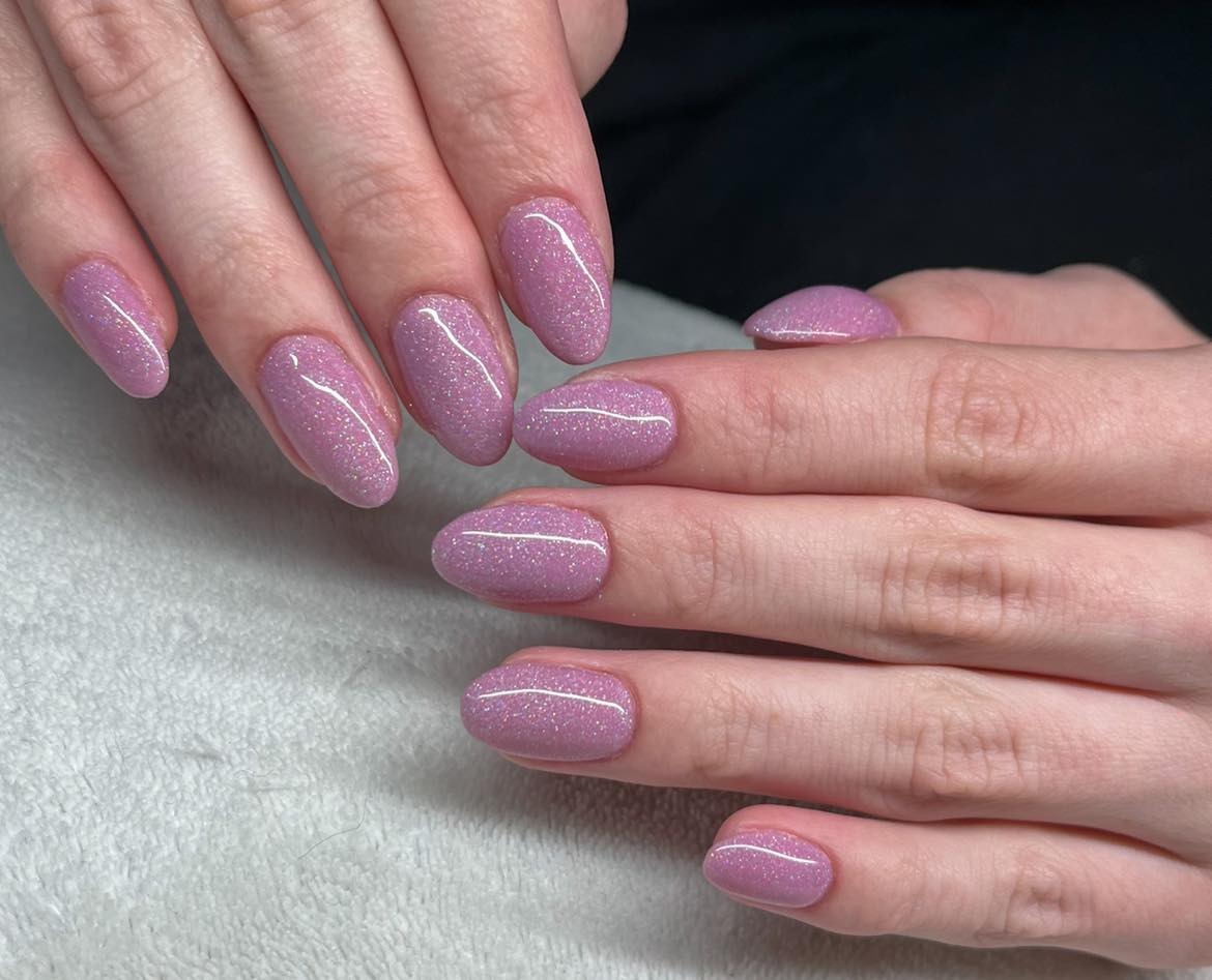 Dłonie z błyszczącym, liliowym manicure hybrydowym w kształcie migdała i owalu, na jasnym, teksturowanym tle.