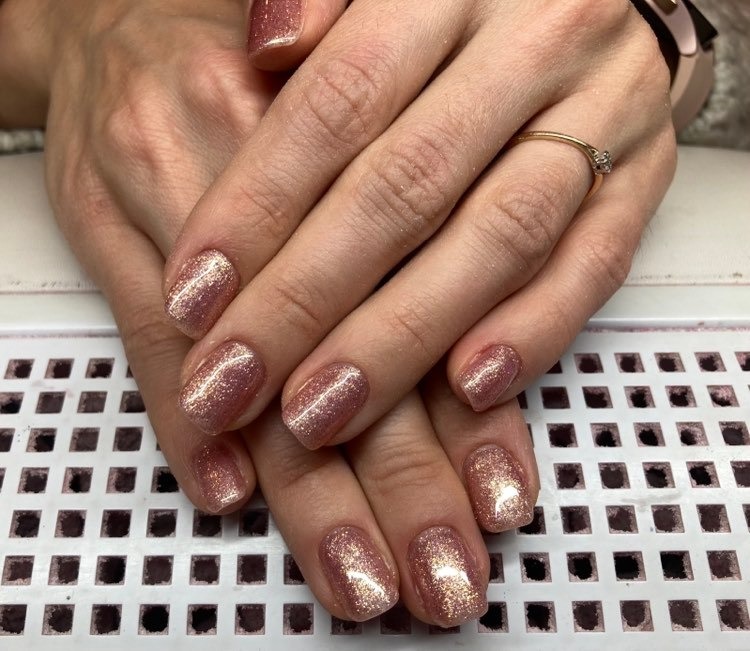 Ręce z błyszczącym, różowo-złotym manicure hybrydowym, wykonanym w Warszawie, na tle kratki lampy UV.