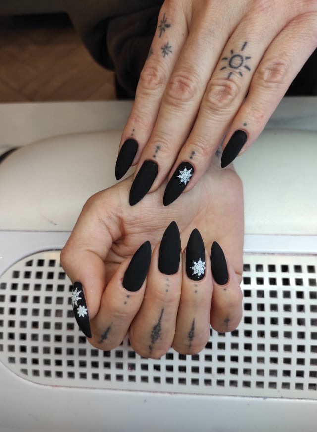 Dłonie z matowymi, czarnymi paznokciami w kształcie szpiców, ozdobione białymi motywami śnieżynek, widoczne tatuaże na palcach.
