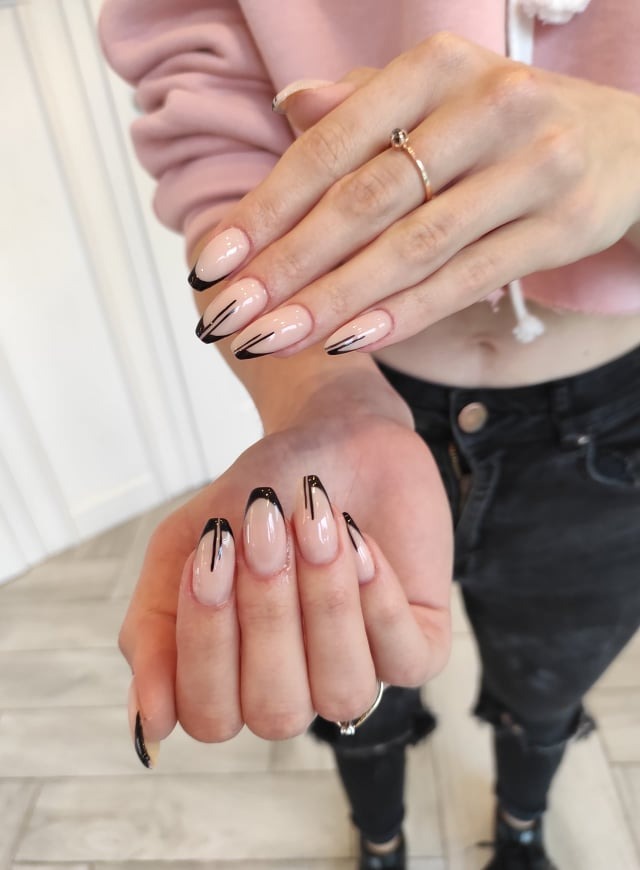 Manicure hybrydowy z beżową bazą i geometrycznym, czarnym zdobieniem na długich paznokciach, widoczne dwie dłonie z pierścionkami