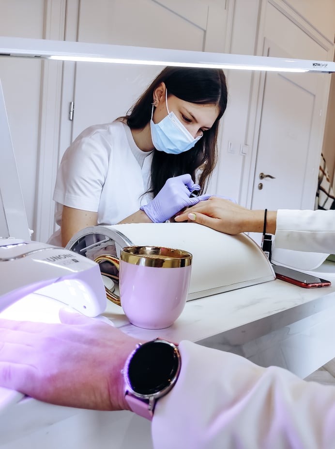 Manikiurzystka w maseczce nakłada lakier na paznokcie klientki, obok lampa UV i różowy kubek z złotym rantem.