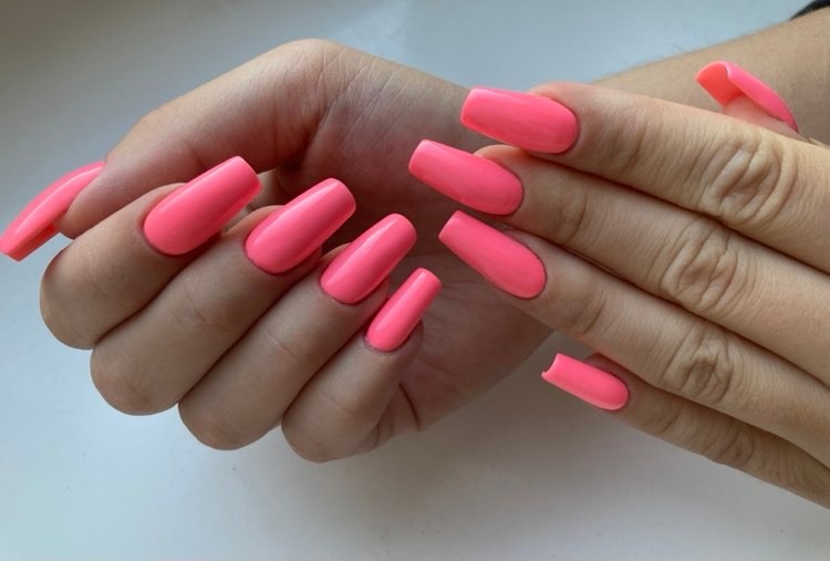 Dłonie z długimi, kwadratowymi paznokciami w neonowym, koralowym odcieniu, ułożone na jasnym tle.