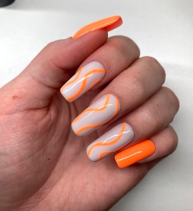 Dłoń z pomalowanymi paznokciami w kolorze neonowej pomarańczy i jasnego beżu, zdobionymi falistymi paskami w odcieniu pomarańczowym.