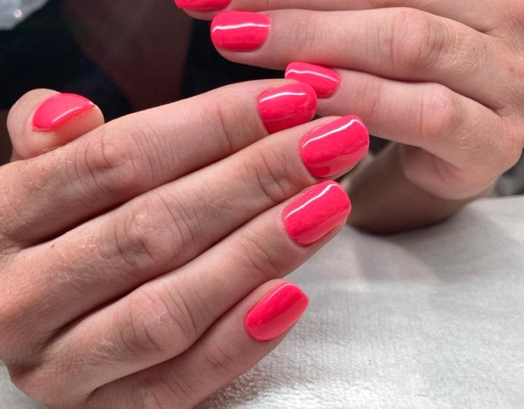 Dłonie z pomalowanymi na jaskrawy róż paznokciami o krótkim, kwadratowym kształcie, widoczne z bliska na jasnym tle.