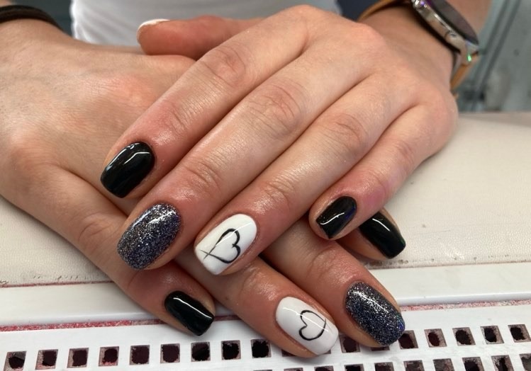 Dłonie z manicure hybrydowym: czarne, brokatowe i białe paznokcie z ręcznie rysowanymi czarnymi sercami, widoczny zegarek na nadgarstku.