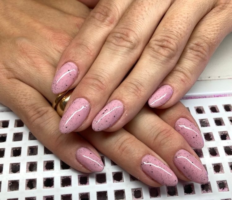 Dłonie z manicure hybrydowym w odcieniu pudrowego różu z czarnymi drobinkami, złoty pierścionek na palcu serdecznym.