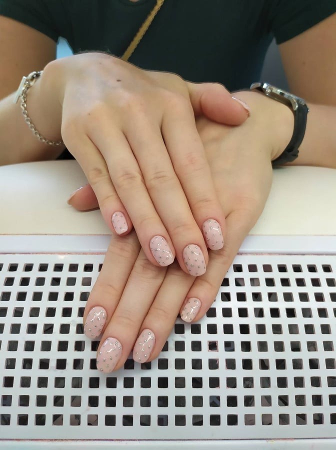 Delikatny manicure hybrydowy w odcieniach beżu z drobnymi srebrnymi zdobieniami na dłoniach kobiety z bransoletką i zegarkiem, ułożonych na białym, perforowanym podłożu.