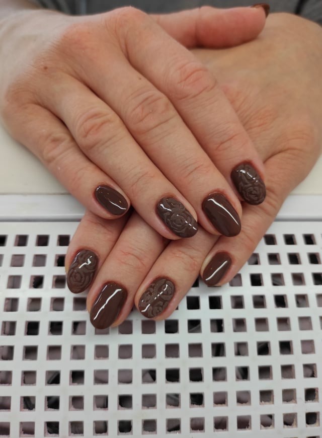 Dłonie z brązowym manicurem hybrydowym, na wybranych paznokciach wytłoczony wzór róży.