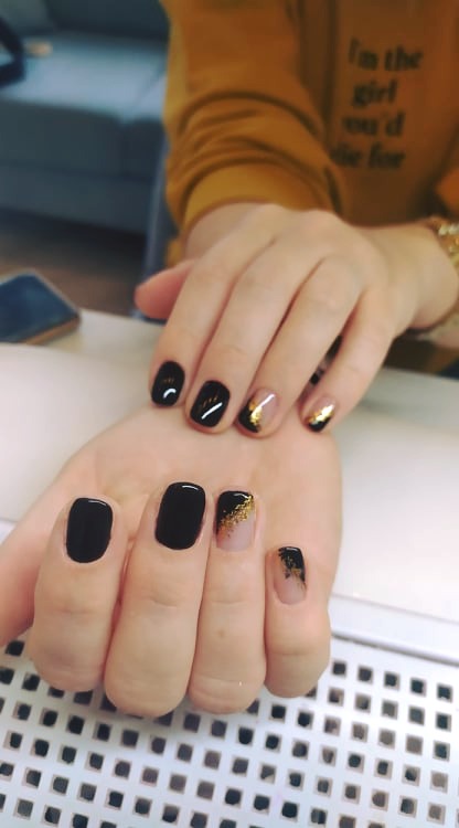 Dłonie z czarnym manicurem i złotymi akcentami na kilku paznokciach, na tle białego stołu i fragmentu żółtej bluzy z napisem.