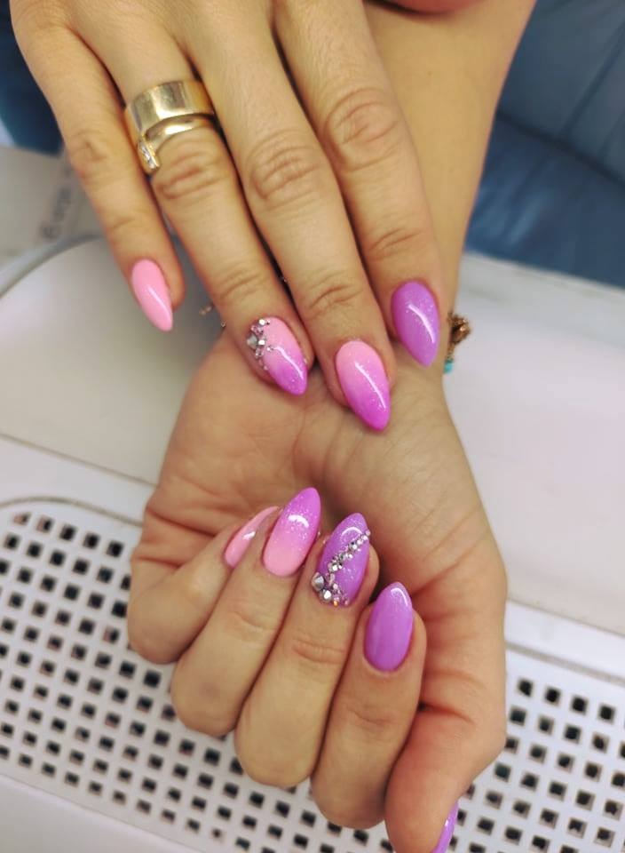 Dłonie z manicure hybrydowym w odcieniach różu i fioletu, z gradientem i drobnymi kryształkami, na tle lampy UV do paznokci.
