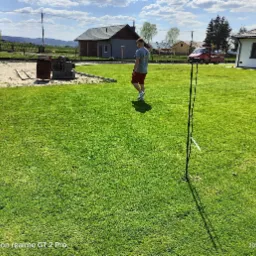 Rozległy, zielony trawnik przed domem jednorodzinnym, z widocznym fragmentem piaskownicy, stertą bloczków betonowych i mężczyzną spacerującym po trawie w słoneczny dzień. W tle widoczny krajobraz...