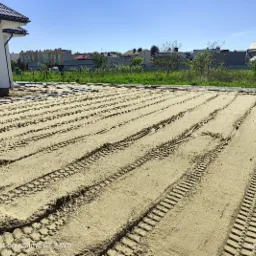 Przygotowany teren pod trawnik, widoczne ślady po maszynach wyrównujących, w tle dom jednorodzinny i zabudowa osiedlowa.