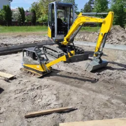 Żółty minikoparka i zagęszczarka gruntu Wacker Neuson na placu budowy z widocznymi fundamentami i przygotowanym podłożem, w tle dom i zieleń.