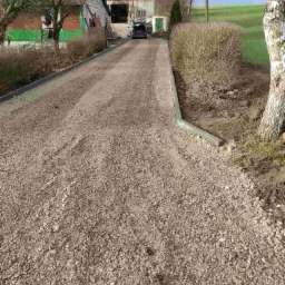 Utwardzona droga dojazdowa do posesji z samochodem w tle, ograniczona zielonymi obrzeżami.