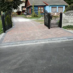 Ułożona z czerwonej kostki brukowej aleja prowadząca do domu z otwartą, kutą bramą, otoczona szarą obwódką z kostki granitowej.