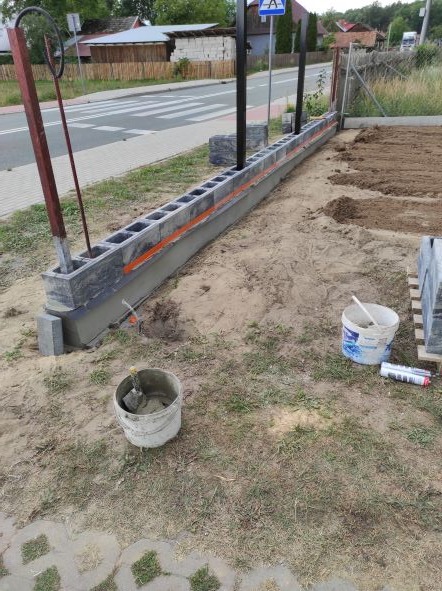 Wznoszenie murku z betonowych bloczków szalunkowych, widoczna warstwa zaprawy, poziomica, wiaderka i narzędzia na placu budowy, w tle ulica i znak drogowy.