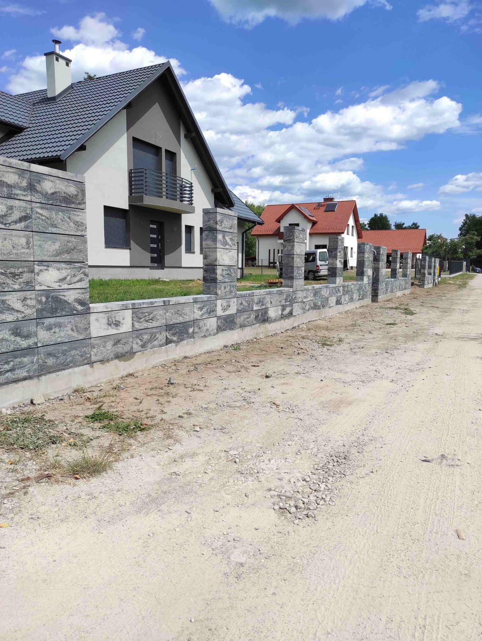 Nowoczesny, szary mur z betonowych bloczków oddzielający posesję od drogi gruntowej. W tle dom z szarą dachówką i fragmenty innych budynków.