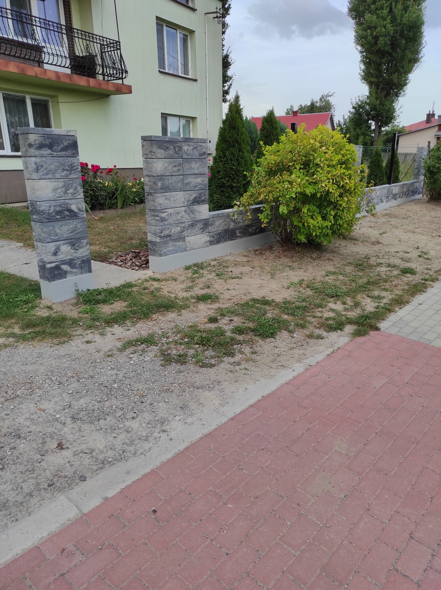 Nowoczesny, szary płot z betonowych bloczków otaczający posesję z trawnikiem i krzewami, widoczny fragment czerwonej kostki brukowej i elewacja domu w tle.