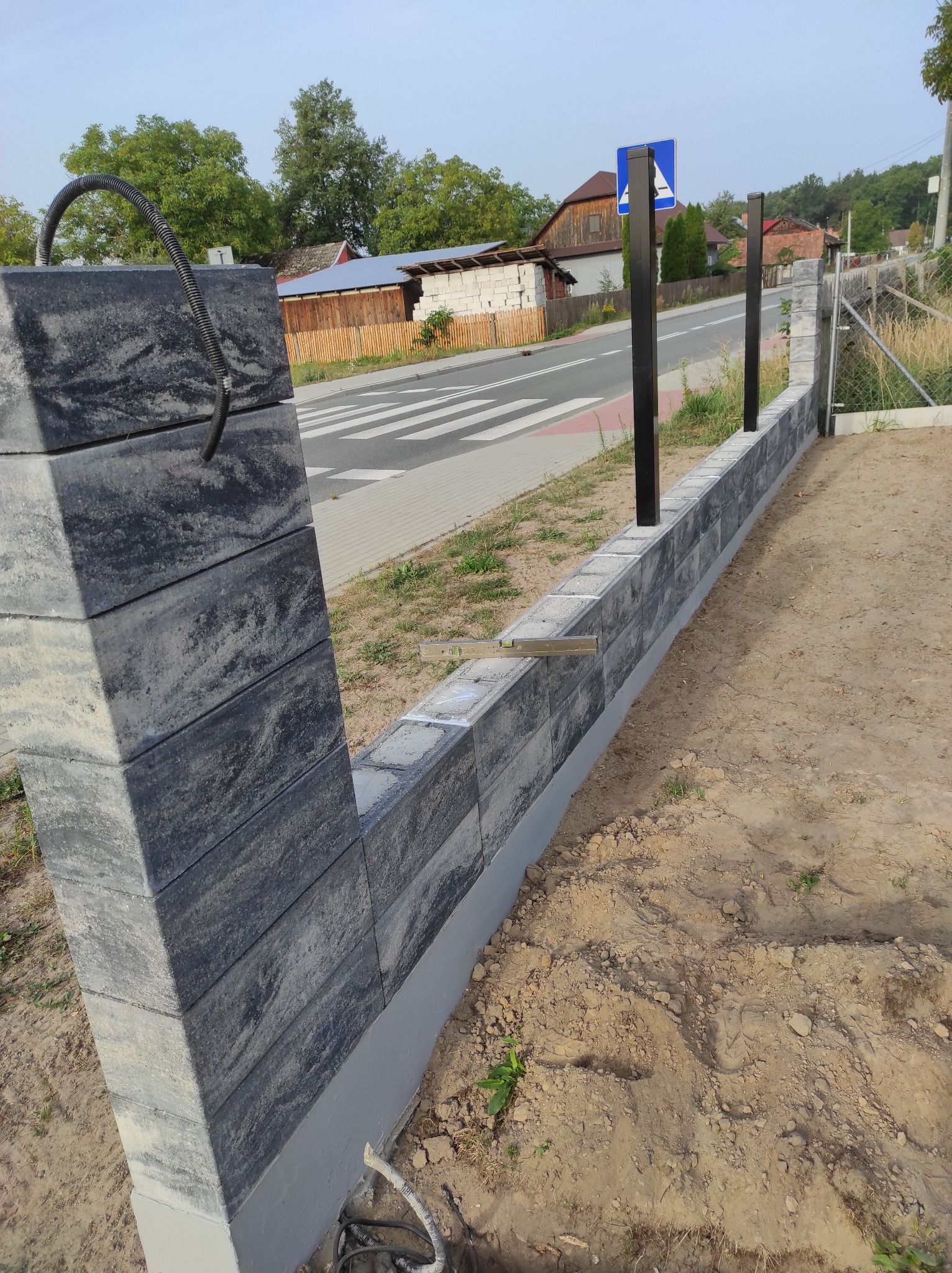 Wznoszony mur z elementów betonowych, z widocznymi czarnymi słupkami ogrodzeniowymi, poziomica na murze, w tle ulica i zabudowania.