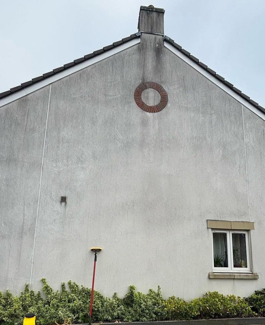 Szara elewacja domu z widocznymi zabrudzeniami i okrągłym elementem dekoracyjnym. Długa szczotka do mycia oparta o ścianę. Widoczne okno i fragment roślinności.