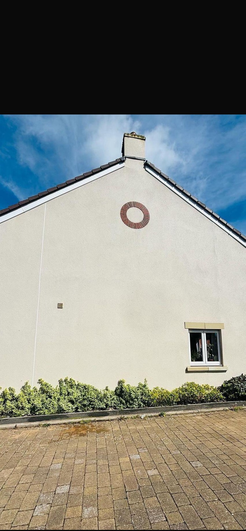 Jasna elewacja domu z ceglanym okrągłym elementem dekoracyjnym, małe okno i pas zieleni u podstawy, widziana z poziomu brukowanego chodnika.