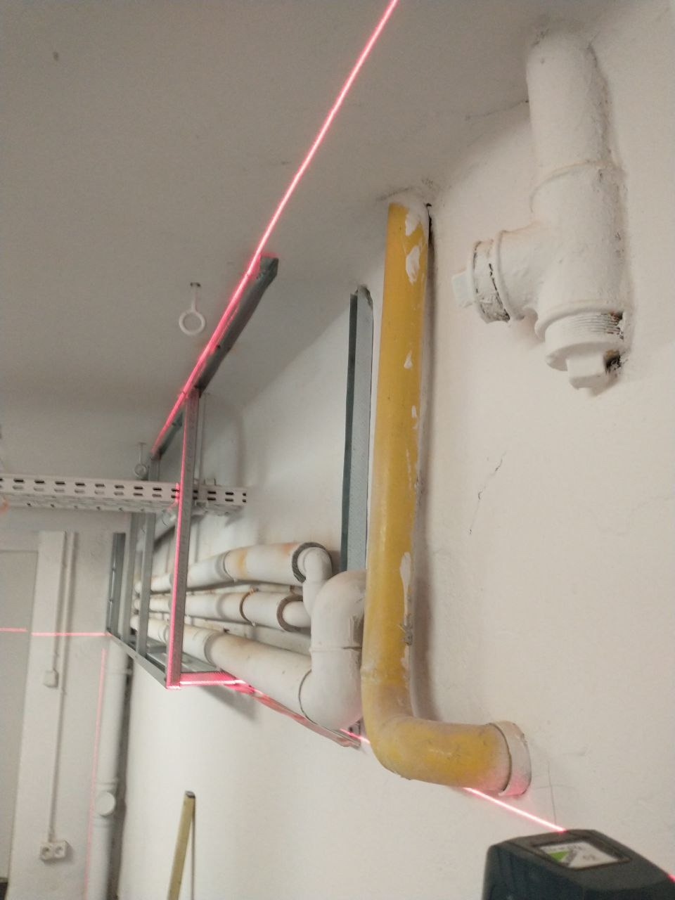 Montaż instalacji pod sufitem z użyciem poziomicy laserowej, widoczne rury i kable w szynach montażowych.