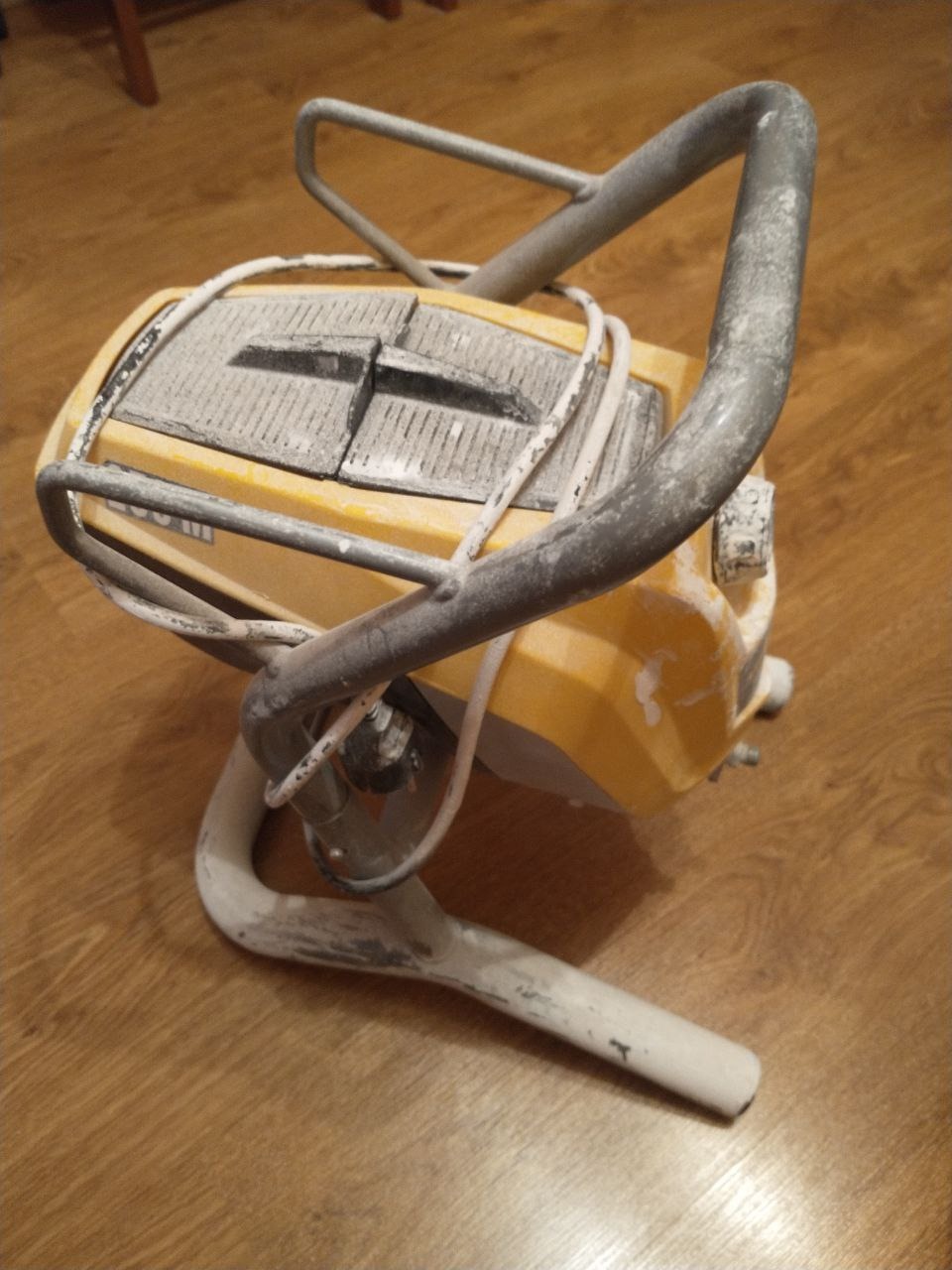 Żółty agregat malarski z metalową ramą, pokryty resztkami farby, leżący na parkiecie.