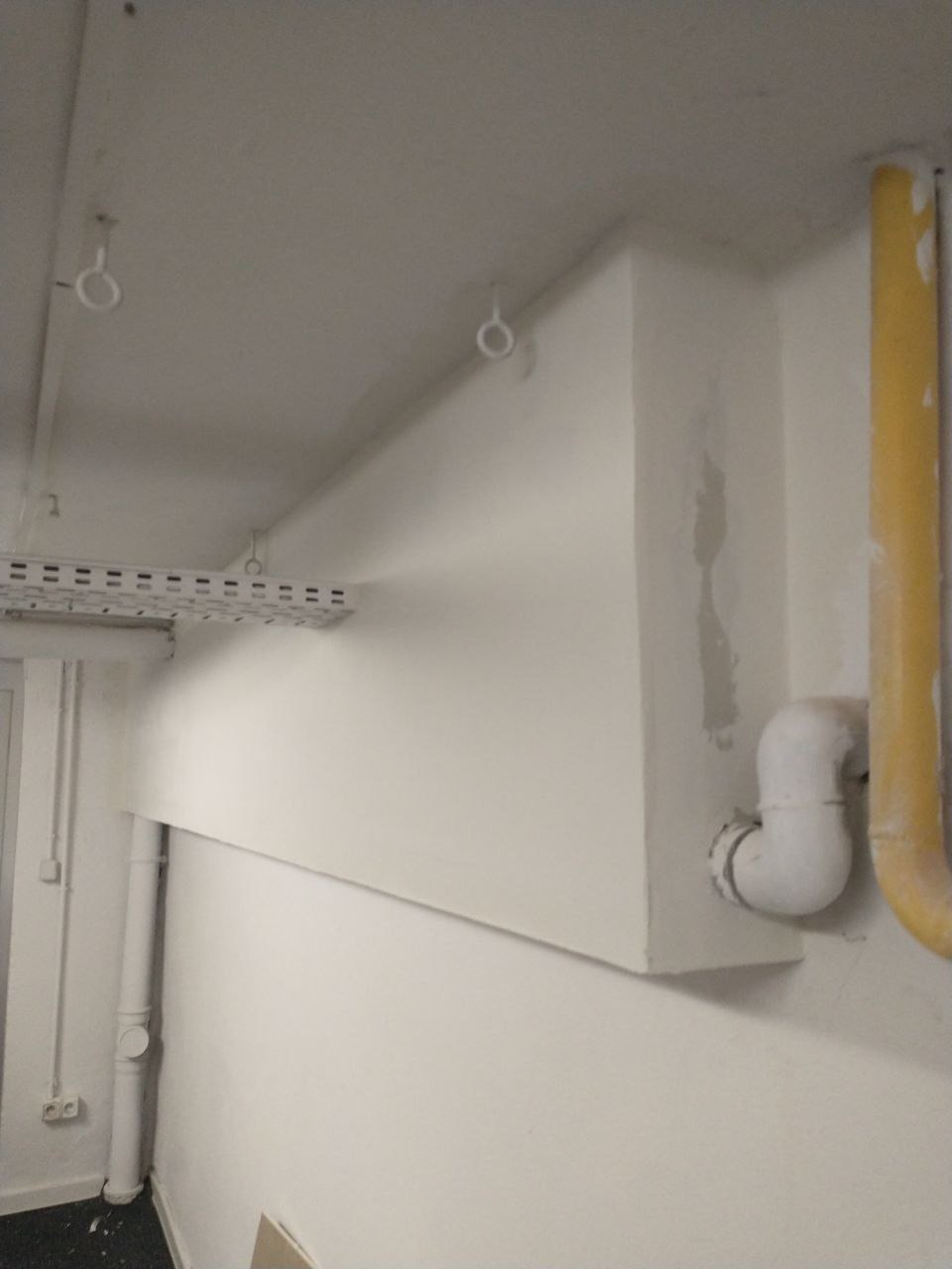Wnętrze budynku z rurami i elementami instalacji pomalowanymi na biało, widoczne ślady po szpachlowaniu na ścianie, żółta rura gazowa w prawym górnym rogu.