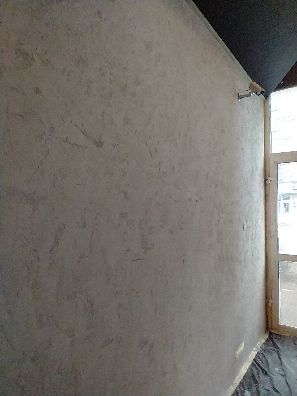 Ściana pokryta szarym mikrocementem z widoczną fakturą, z fragmentem okna i czarnym sufitem w rogu pomieszczenia.