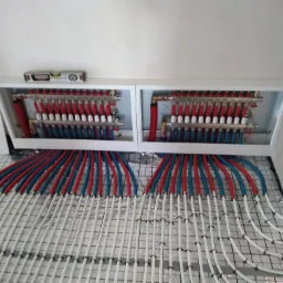 Rozdzielacz ogrzewania podłogowego w białej obudowie, z widocznymi rurami doprowadzającymi wodę w kolorach czerwonym i niebieskim, oraz białymi rurkami grzewczymi ułożonymi na siatce montażowej...