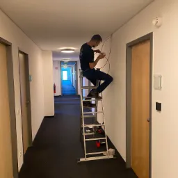 Elektryk na drabinie łączy przewody w korytarzu, widoczny czujnik dymu na ścianie i narzędzia na drabinie.