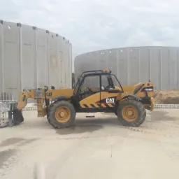 Żółta ładowarka teleskopowa CAT TH360B z platformą roboczą na tle dwóch betonowych zbiorników o cylindrycznym kształcie, widoczne ślady opon na placu budowy.