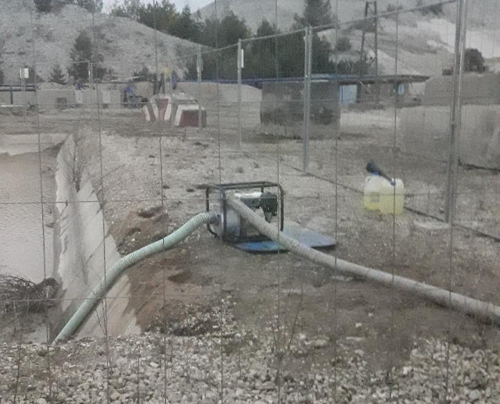 Pompa zanurzeniowa odwadniająca wykop, widoczna siatka ogrodzeniowa, w tle hałda ziemi i pracujący ludzie.