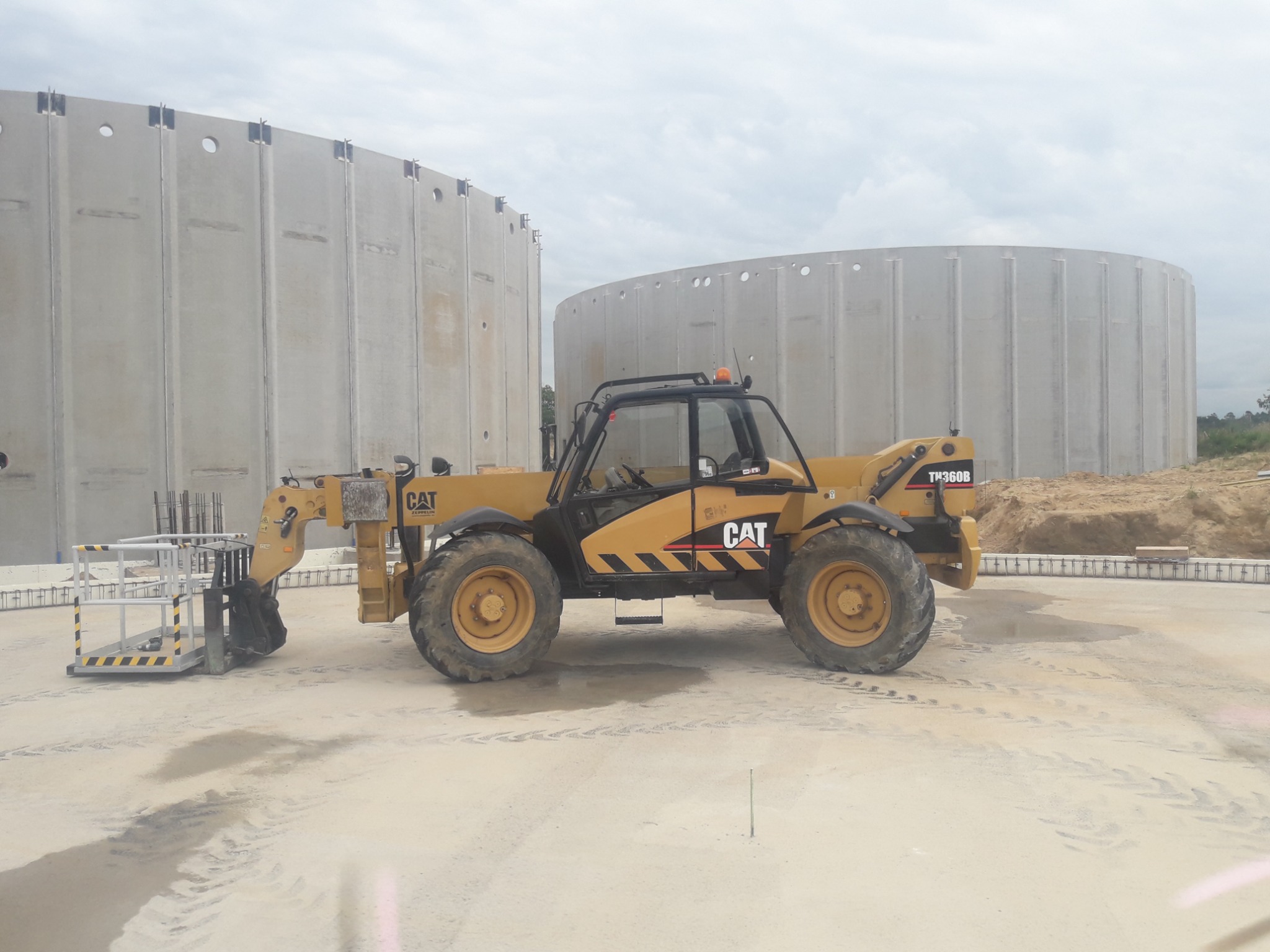 Żółta ładowarka teleskopowa CAT TH360B z platformą roboczą na tle dwóch betonowych zbiorników o cylindrycznym kształcie, widoczne ślady opon na placu budowy.