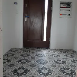 Ułożona podłoga z wzorzystych, szaro-czarnych płytek w przedpokoju z brązowymi drzwiami i białą ścianą z zamontowaną skrzynką elektryczną.