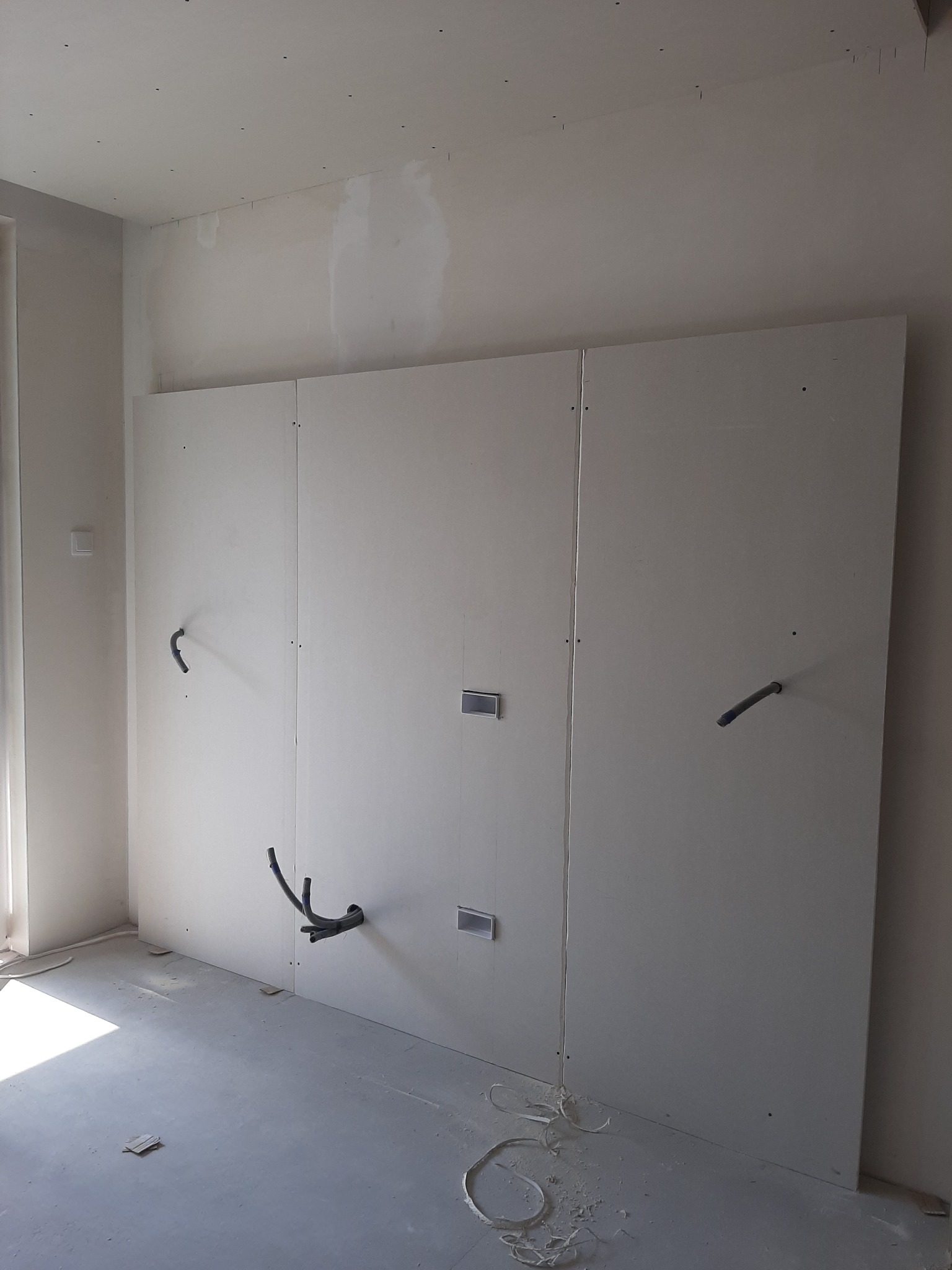 Ściana z płyt kartonowo-gipsowych w trakcie montażu z widocznymi przewodami instalacji elektrycznej i fragmentami instalacji wodnej, surowy beton na podłodze, widoczne łączenia płyt.