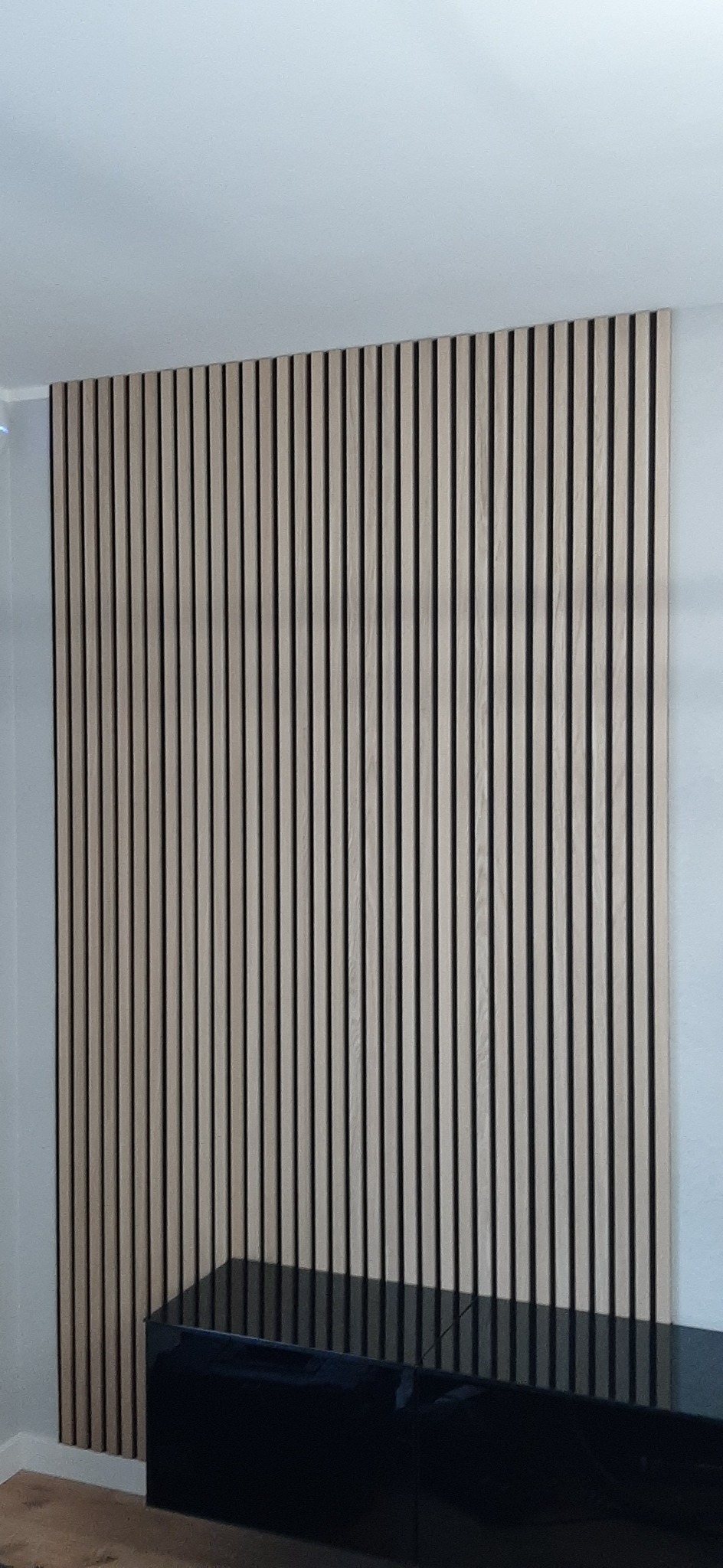 Panel ścienny z drewnianych lameli w jasnym odcieniu, umieszczony za czarną, błyszczącą komodą w nowoczesnym wnętrzu.